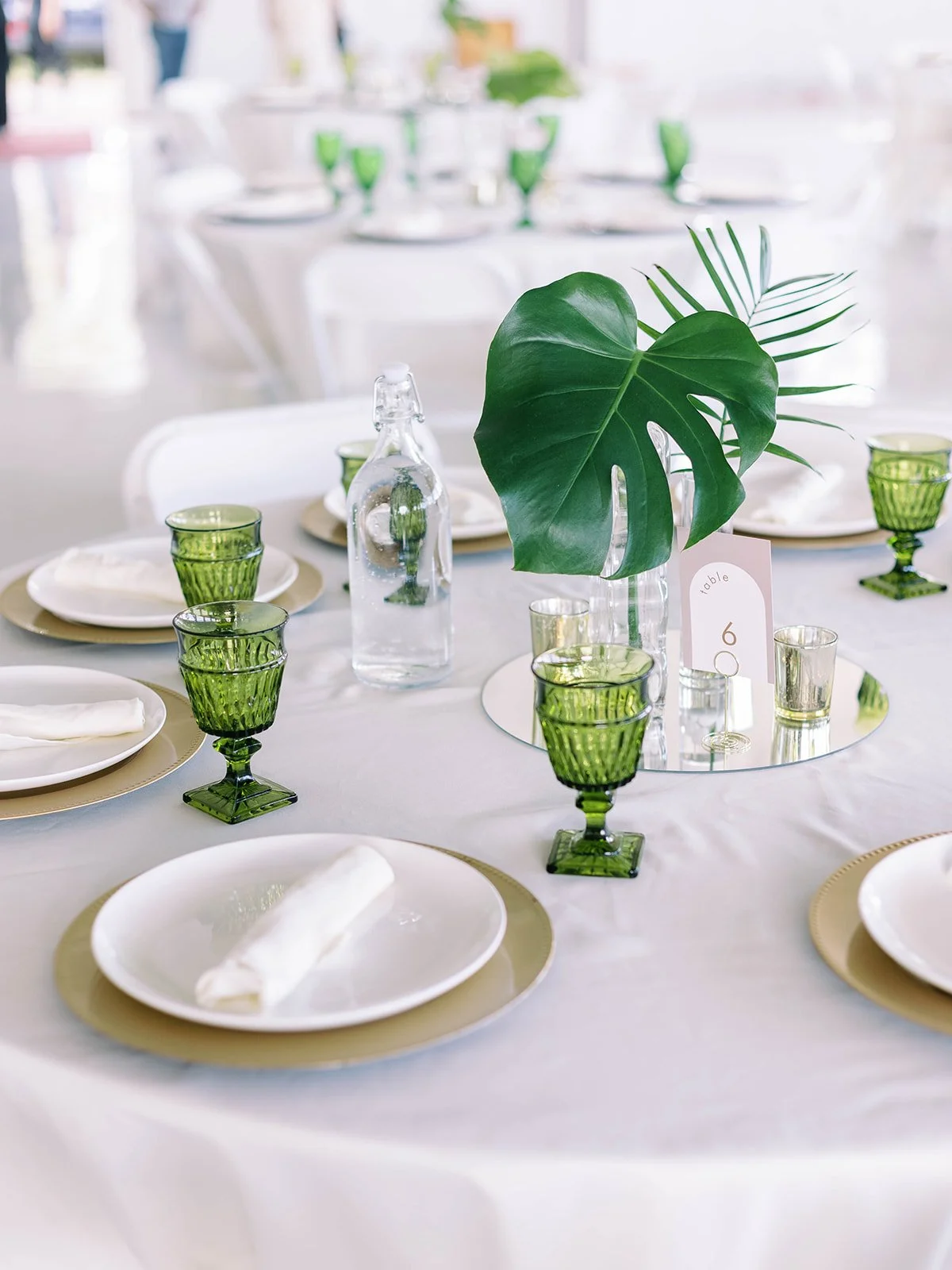 Wedding reception tablescape with green goblets, gold charger plates, white napkins, and a monstera leaf centerpiece on a mirror base with a table number card
