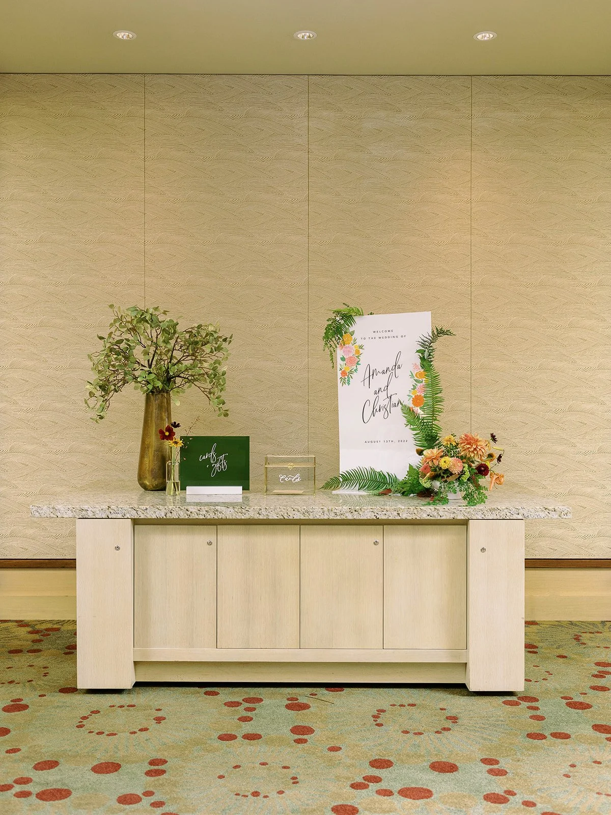 Wide shot of the hotel wedding welcome table featuring the Amanda and Christian sign, a gold vase with greenery, a cards and gifts sign, and a colorful floral arrangement