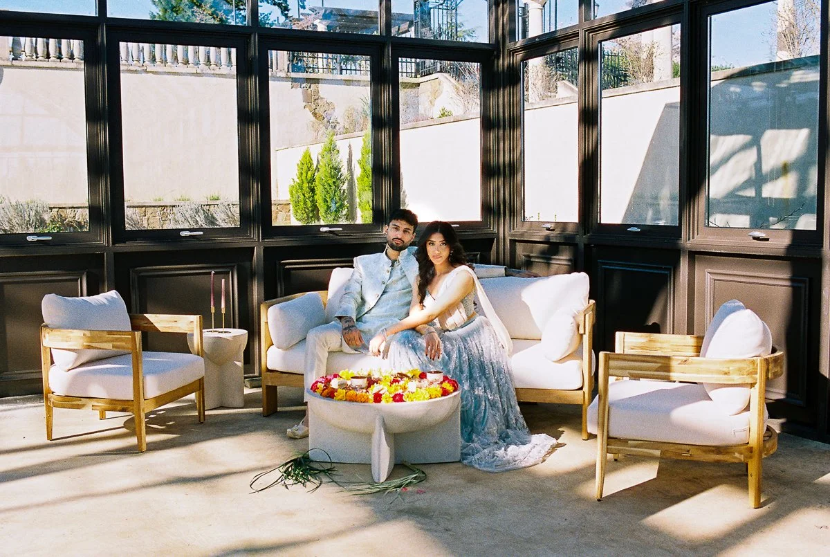 South Asian couple seated in modern lounge setup beside colorful floral arrangement inside conservatory venue.