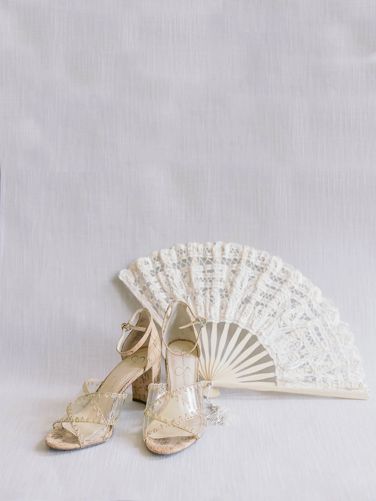 Jessica Simpson cork-heeled sandals with clear straps displayed against a white lace hand fan on a white linen background
