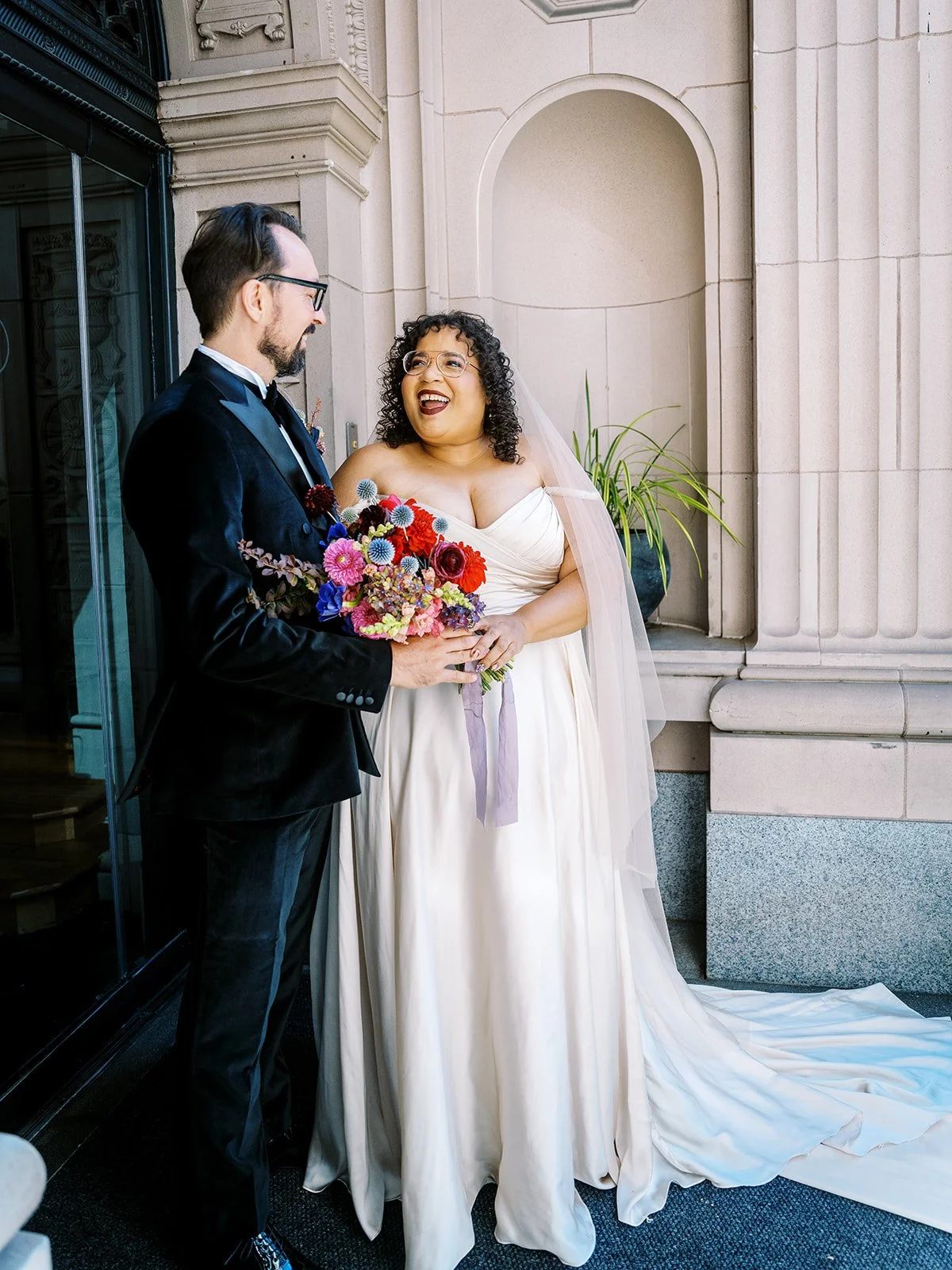 Bride and groom standing at Sentinel Hotel entrance, smiling and holding a colorful bouquet.