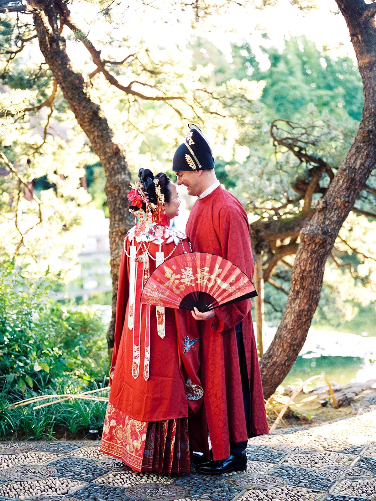 Bride and groom photographed through garden foliage beside the koi pond, both in red traditional attire and smiling at each other
