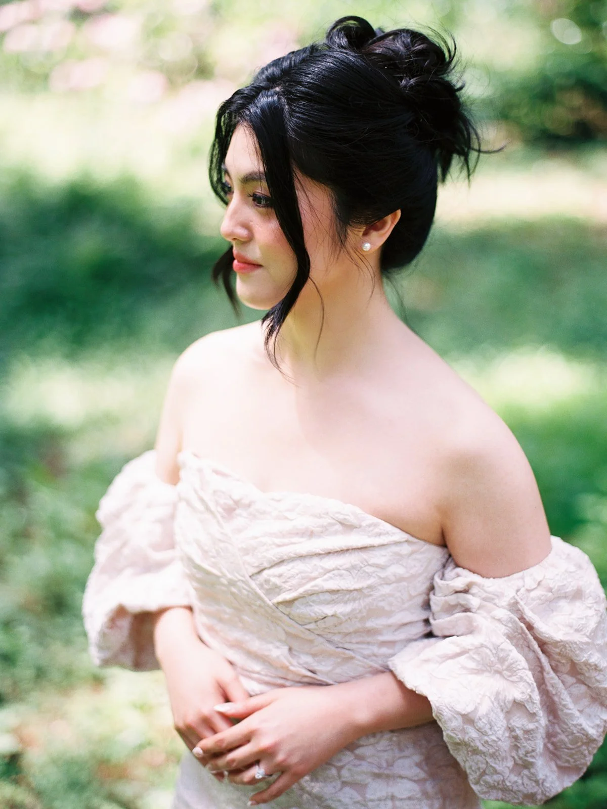 Profile portrait of the bride in a blush off-shoulder gown gazing to the side in soft dappled garden light, pearl earring and engagement ring visible