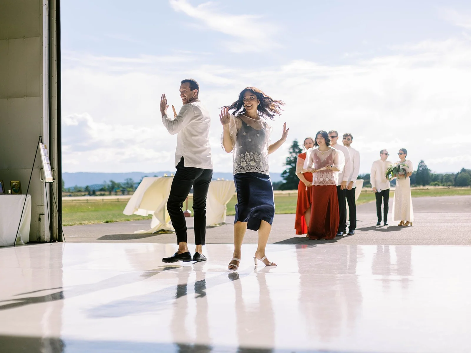 Wedding party member in a navy dress dancing energetically into the Pearson Air Museum hangar during the reception grand entrance, with other party members queued on the tarmac behind
