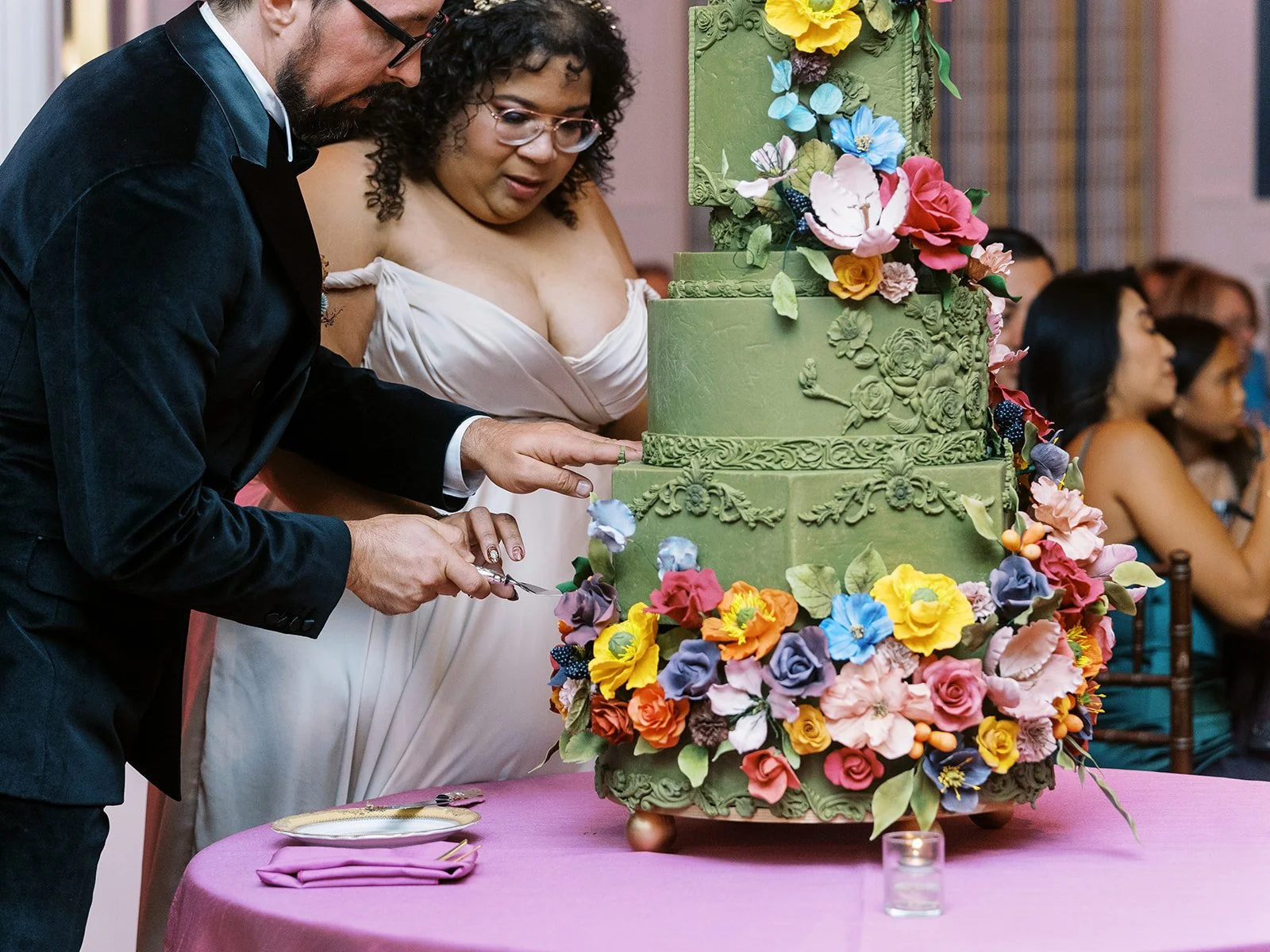 Bride and groom cutting a tall green wedding cake decorated with colorful flowers on a pink tablecloth.