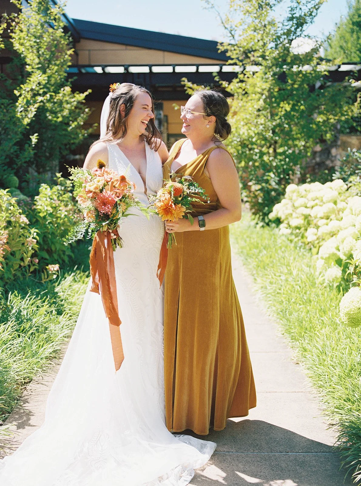 Bride and bridesmaid smiling at each other holding bouquets on sunlit path