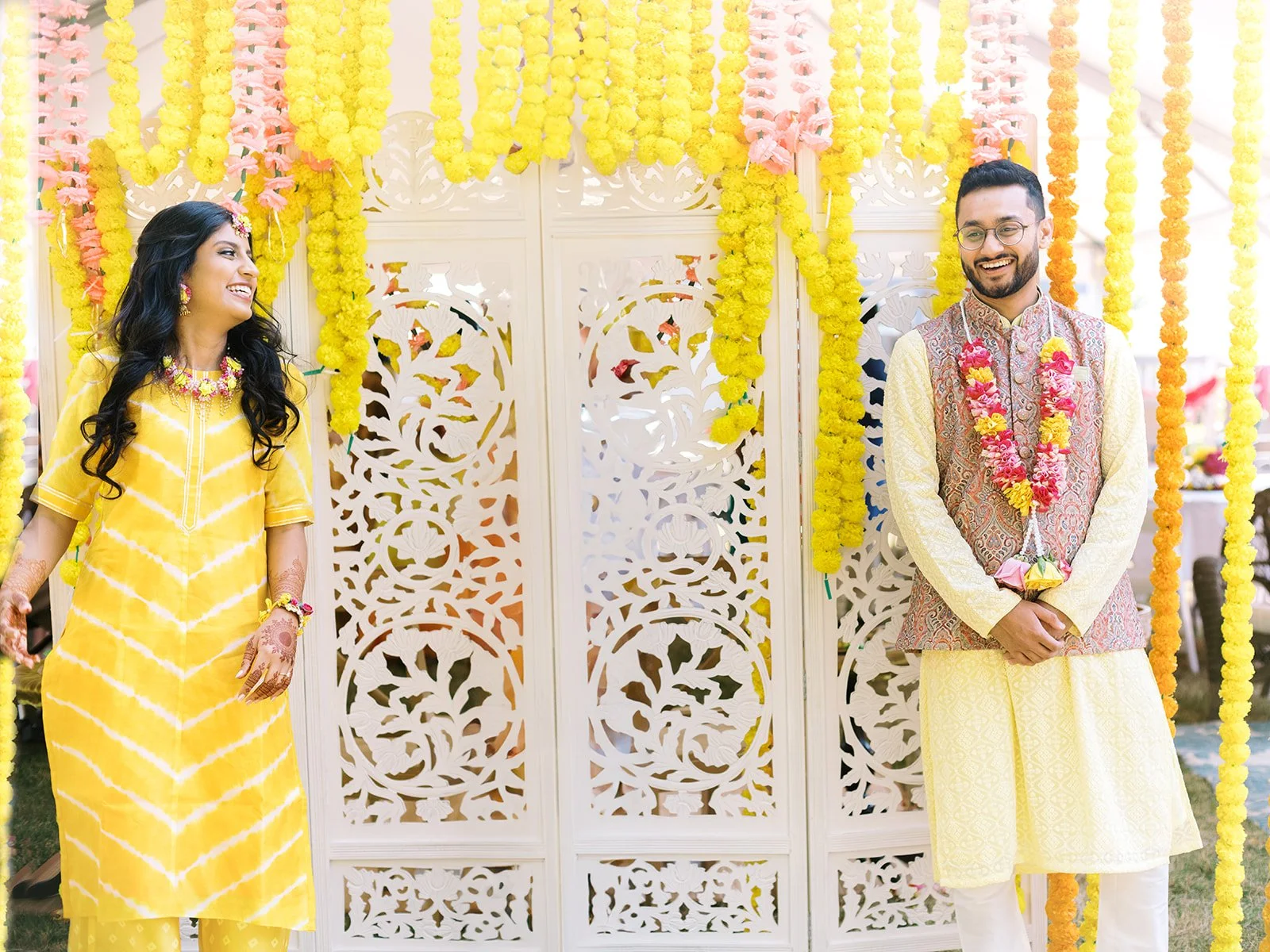 Couple in yellow traditional Indian outfits smiling at each other beneath marigold garlands during a Haldi ceremony.