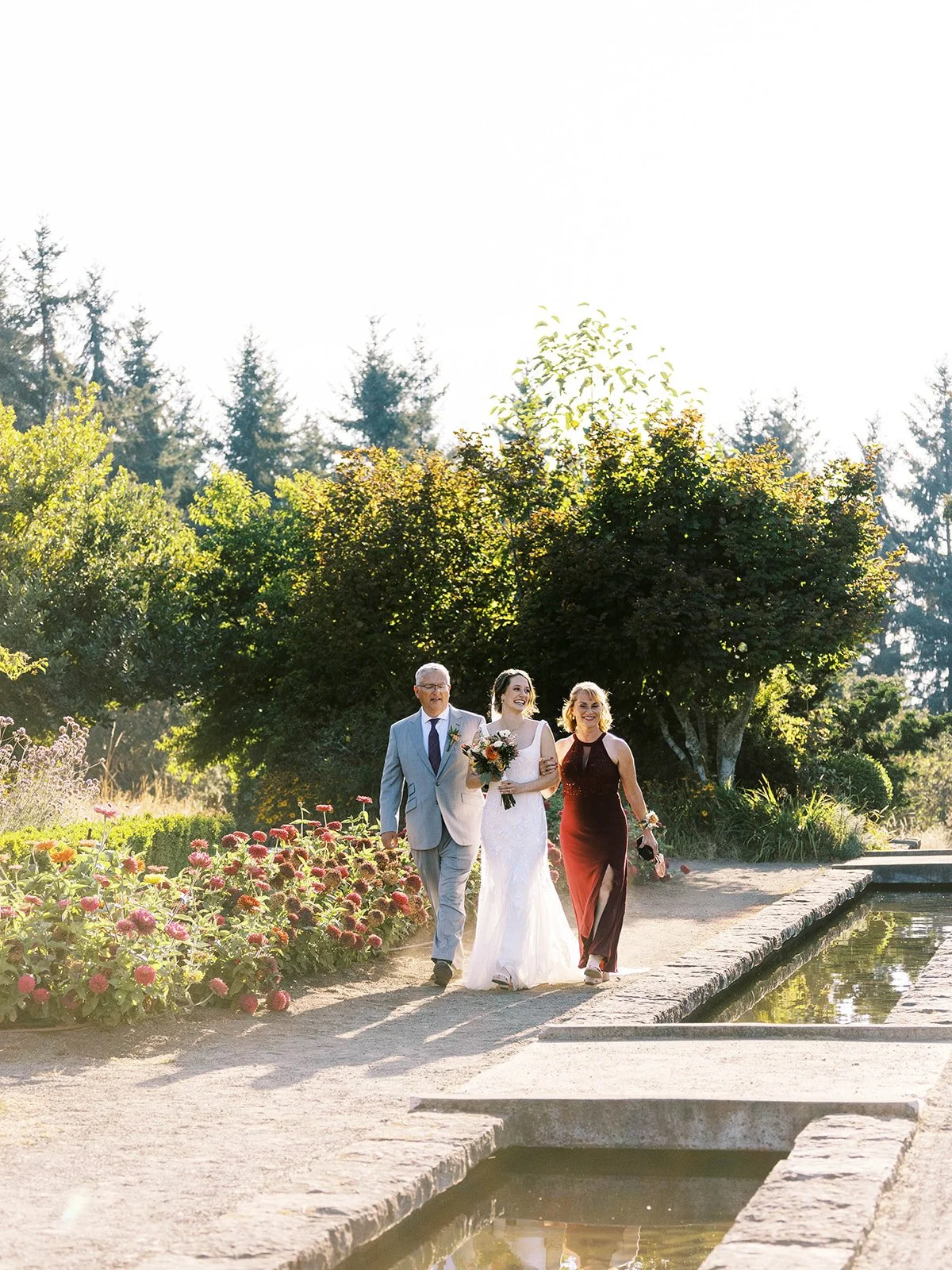 Bride walking with parents along garden path, holding bouquet near water feature