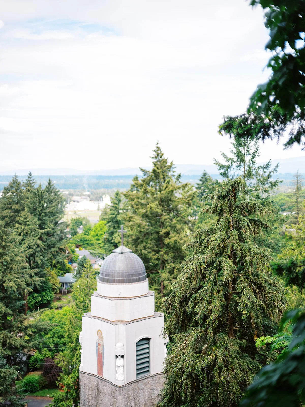 Elevated view of The Grotto Portland's domed bell tower with a cross, surrounded by towering evergreens and overlooking a sweeping view of Northeast Portland