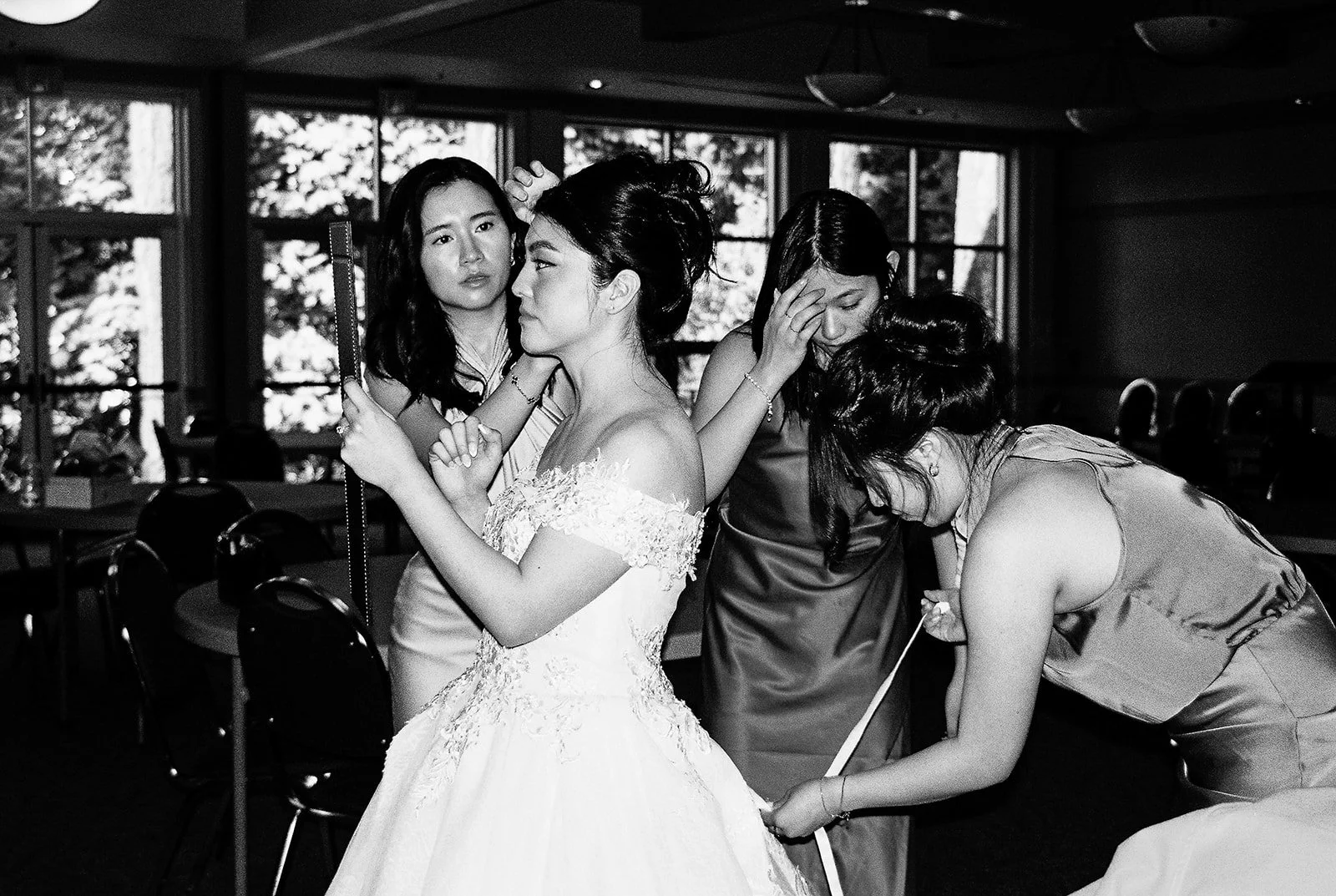 Black and white photo of bridesmaids in satin dresses fussing over the bride — fixing her hair and gown — inside The Grotto's reception hall