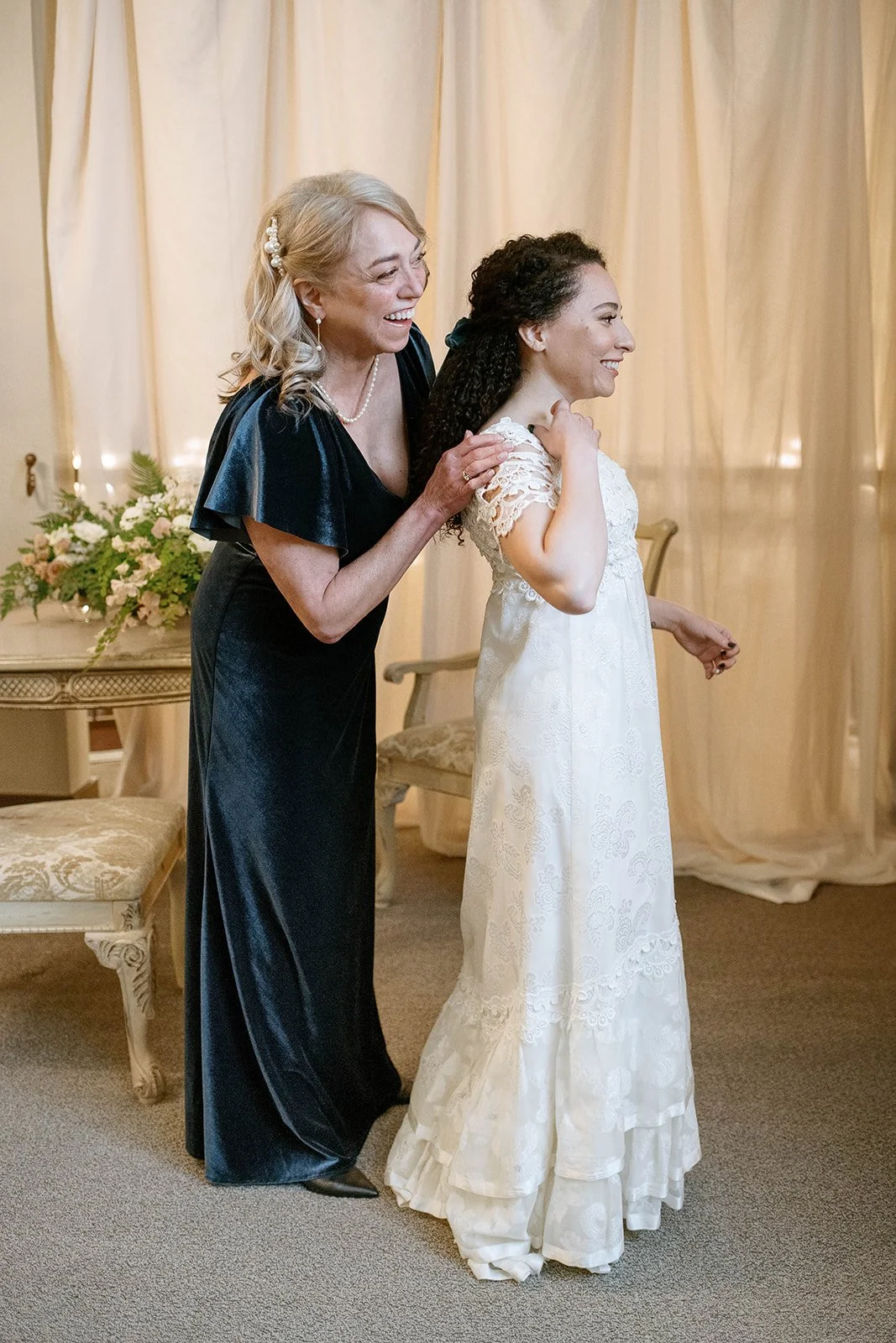 Bride in her vintage lace wedding gown as a woman helps adjust the dress before the ceremony.
