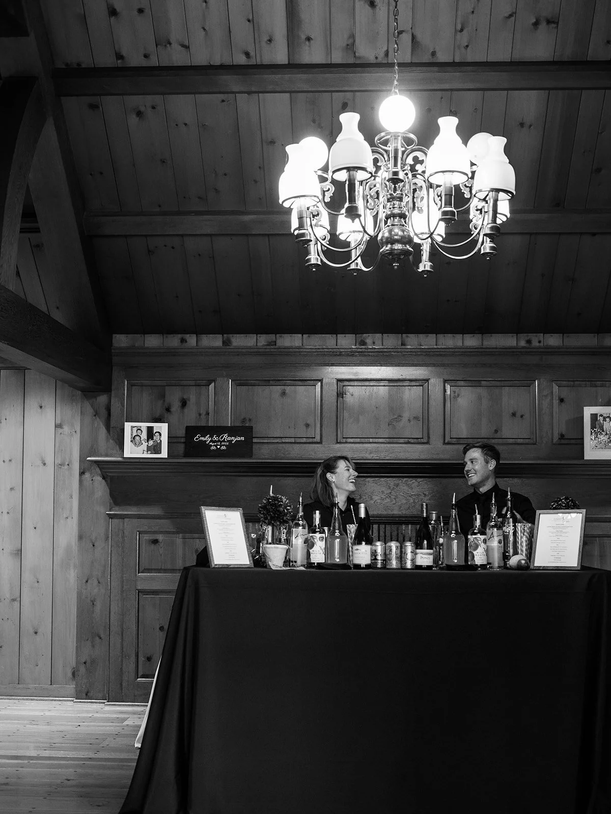 Black-and-white scene of two wedding guests seated behind a bar beneath a chandelier.