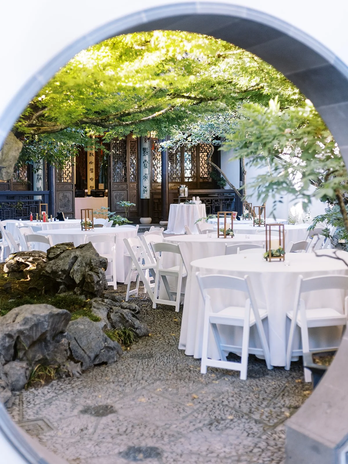 The wedding reception courtyard viewed through a circular moon gate, round tables with white linens and wood lantern centerpieces set among scholar's rocks and a canopy of green trees.