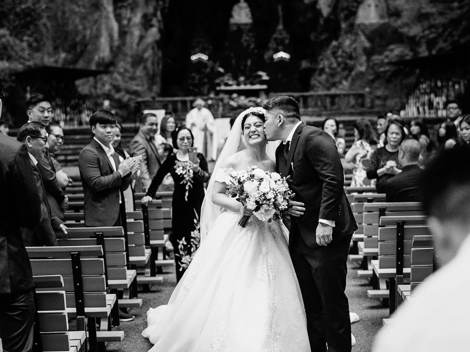 Black and white photo of the groom kissing the bride's temple as they recess up the aisle at The Grotto, guests applauding and smiling on both sides