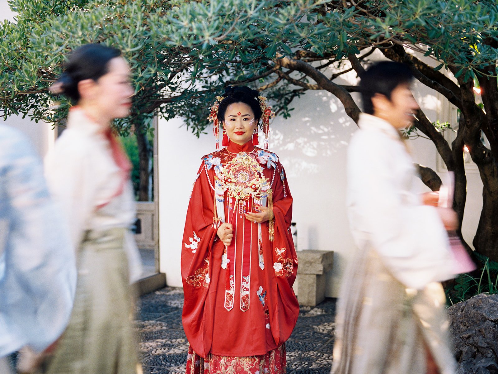 Bride in red qun kwa standing still among blurred attendants in motion beneath a garden tree at Lan Su Chinese Garden