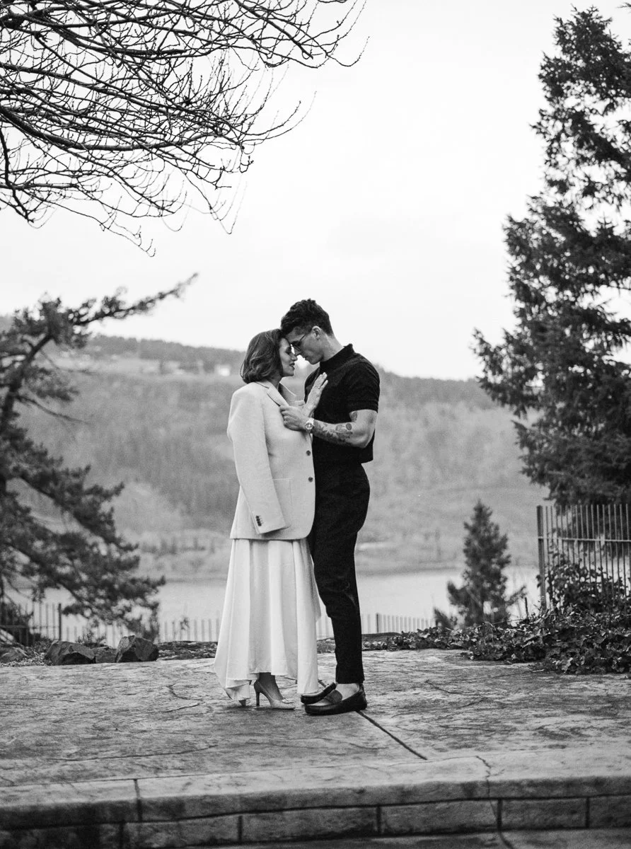 Black and white portrait of couple standing close outdoors, foreheads touching with lake and trees in background