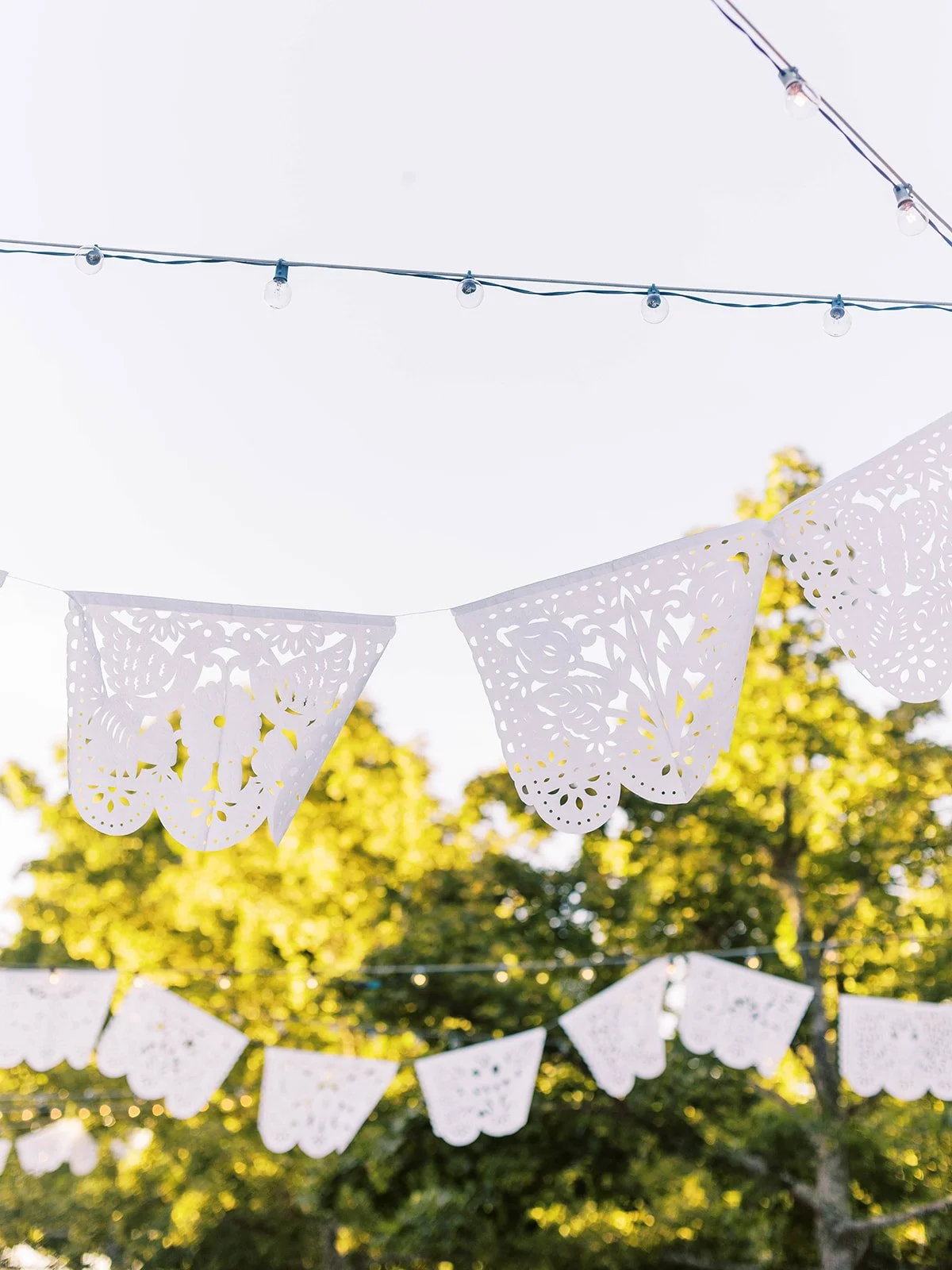 White papel picado banners and string lights hanging above an outdoor garden wedding reception.