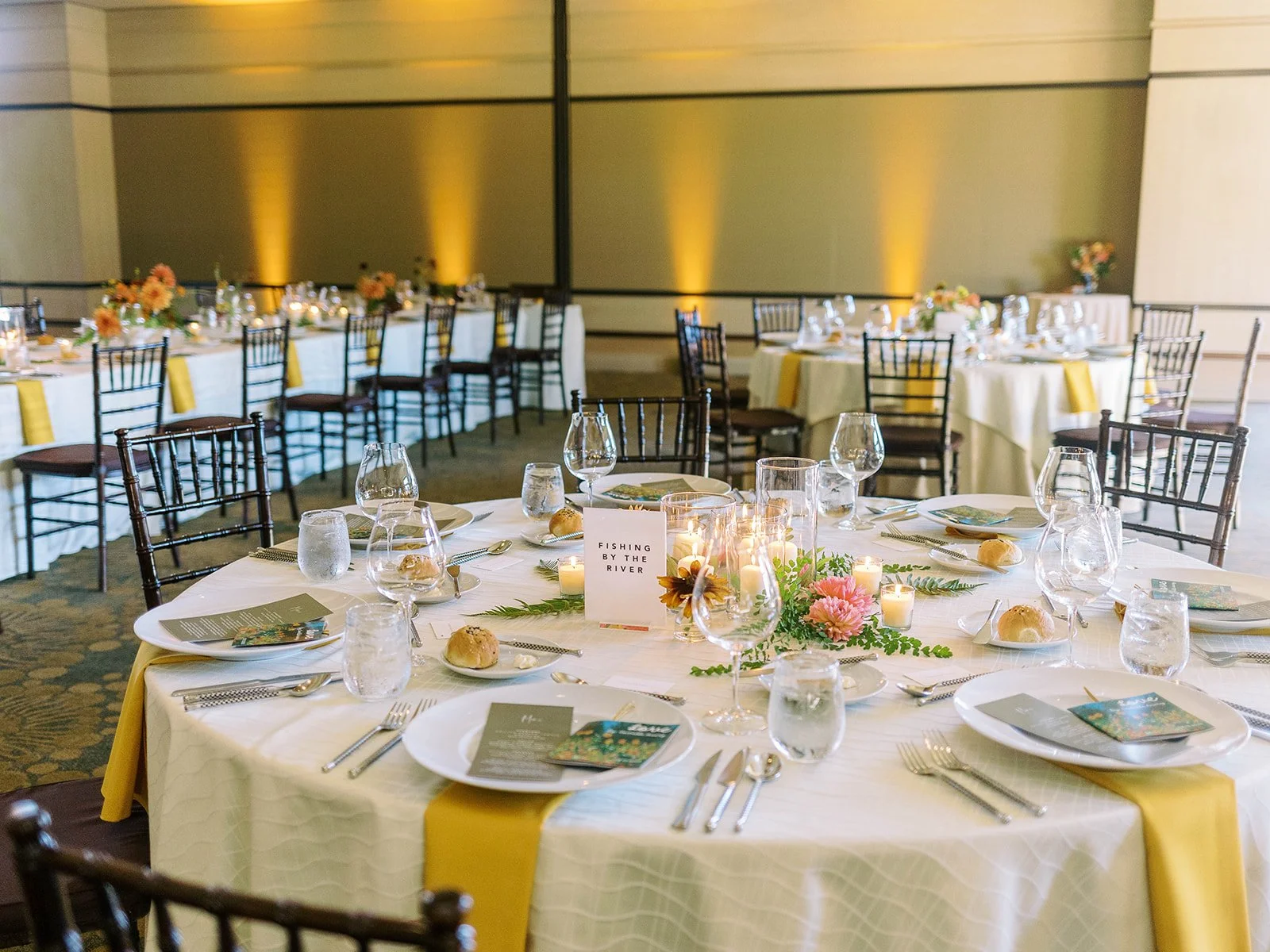Round reception table set with white linens, yellow napkins, candles, pink floral and greenery centerpiece, and a table card reading "Fishing by the River," with the full amber-lit ballroom behind it