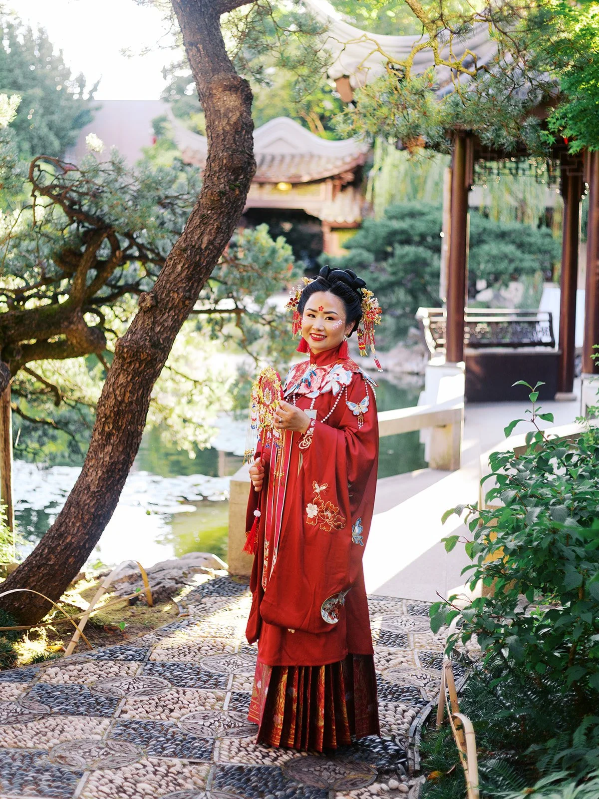 Bride in full red qun kwa and traditional hair ornaments standing alone on a pebbled garden path beside the koi pond at Lan Su Chinese Garden, a pavilion visible behind her