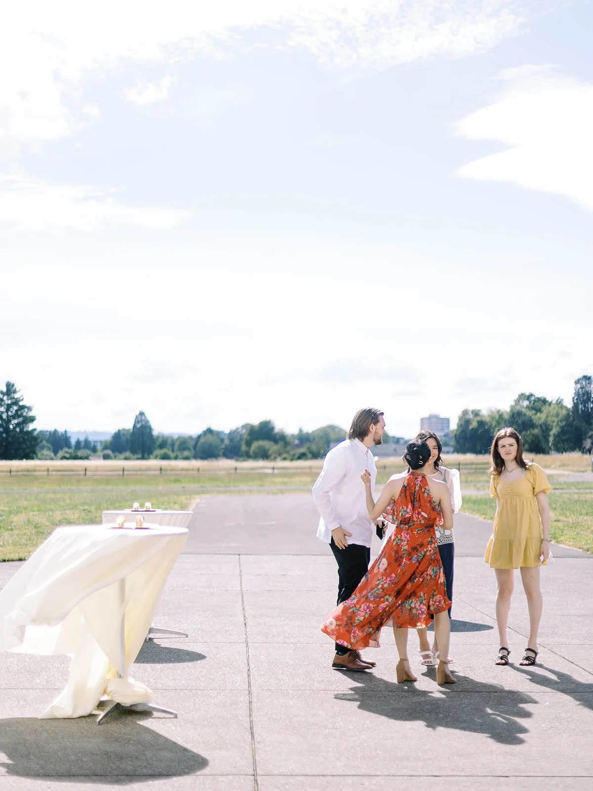 Three wedding guests mingling on the sunny Pearson Airfield tarmac beside a draped cocktail table
