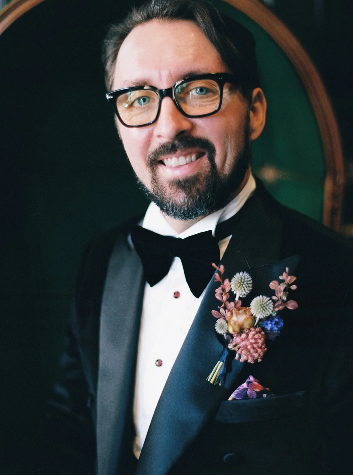 Close-up of groom wearing glasses and black tuxedo with floral boutonniere.
