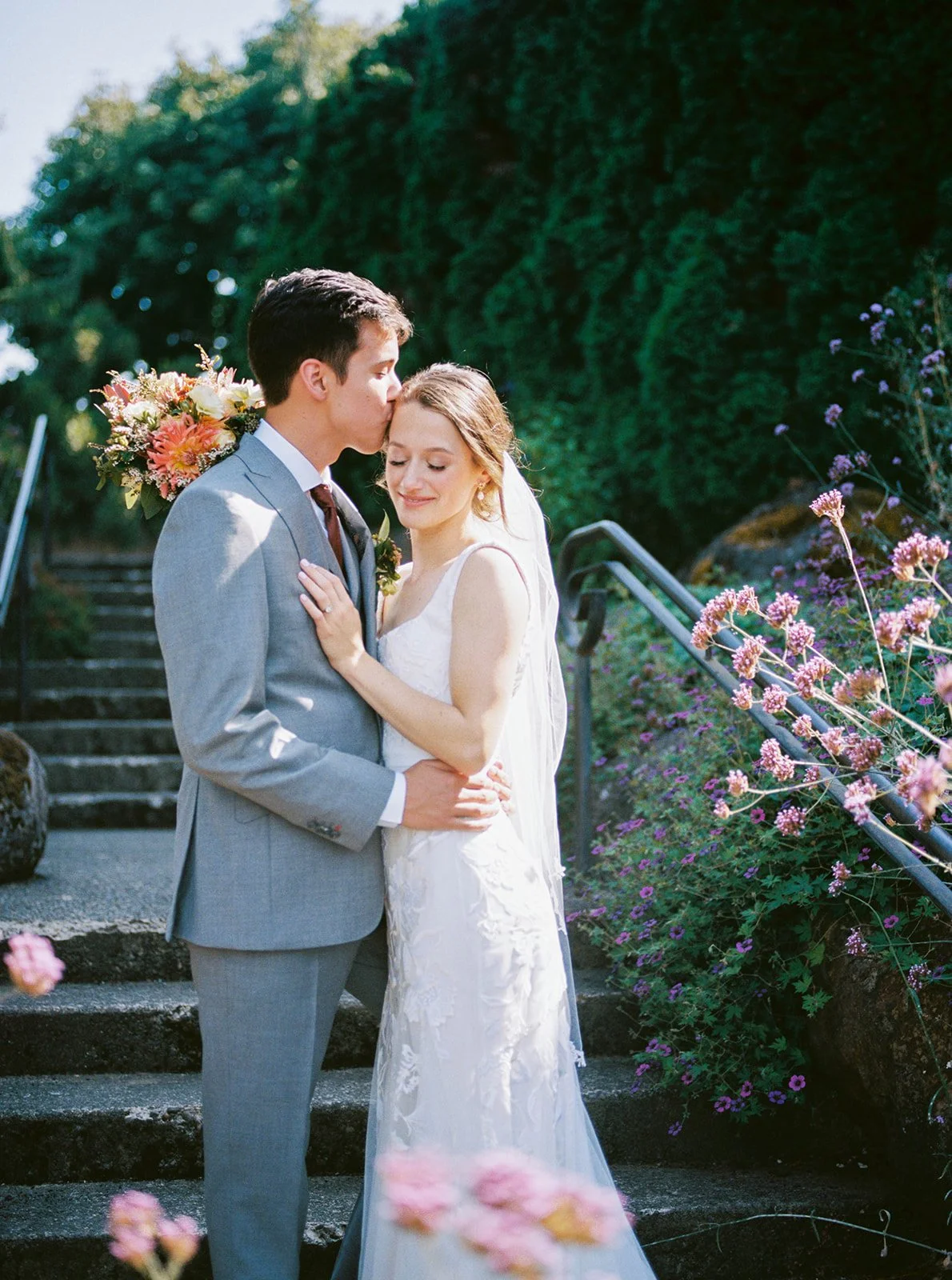 Groom kisses bride’s forehead as they embrace on stone steps lined with flowers at Oregon Gardens.
