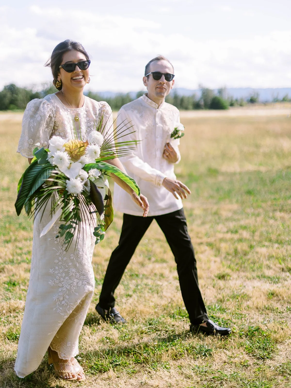 Bride laughing while walking through a sunny field carrying a tropical bouquet, groom following behind in barong tagalog and sunglasses
