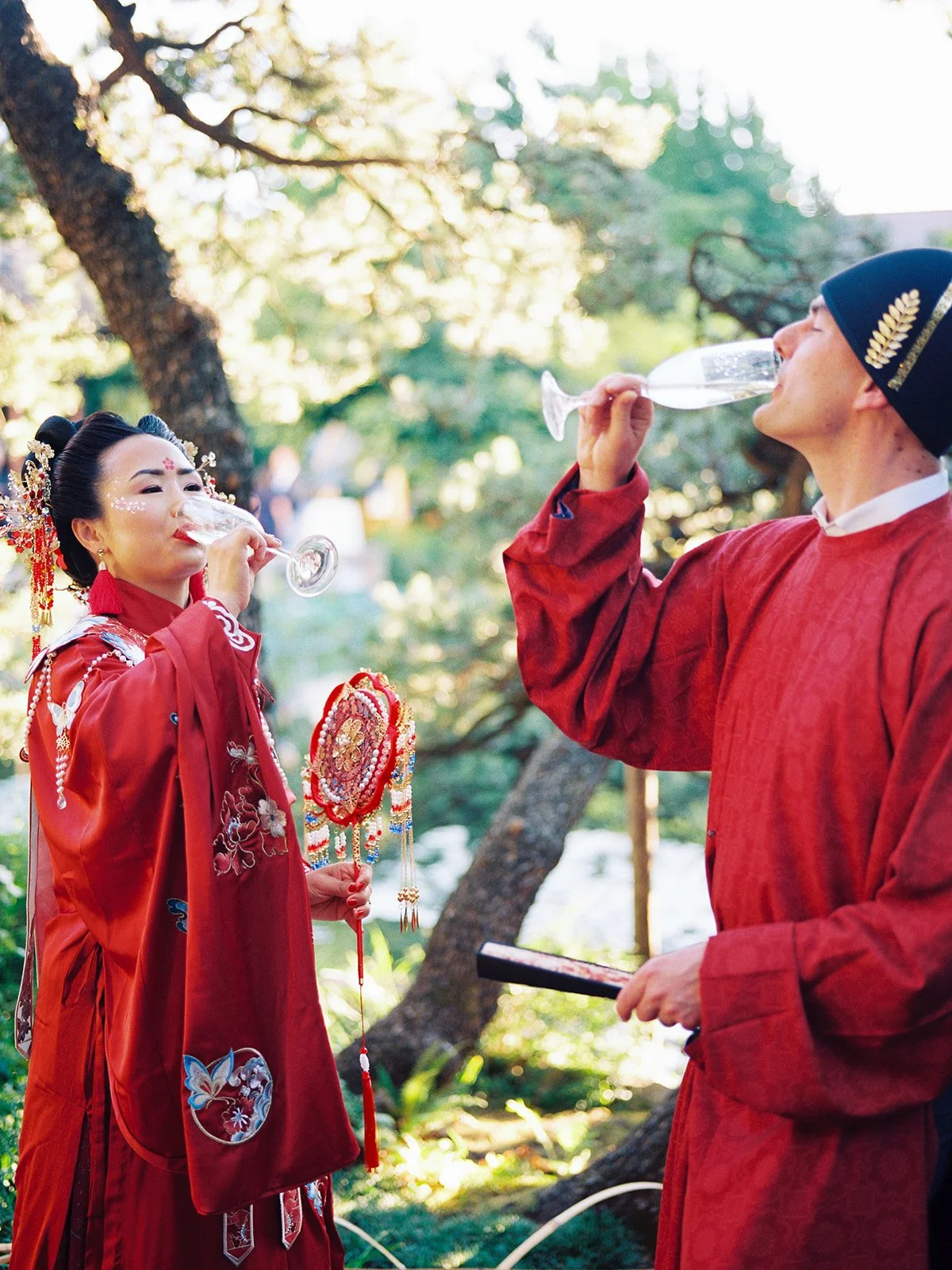 Bride and groom in full traditional red Chinese wedding attire toasting with champagne flutes beneath a pine tree at Lan Su Chinese Garden