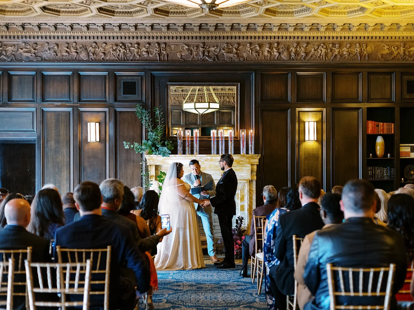 Wide view of indoor wedding ceremony with couple at fireplace altar and guests seated in gold chairs beneath ornate ceiling.