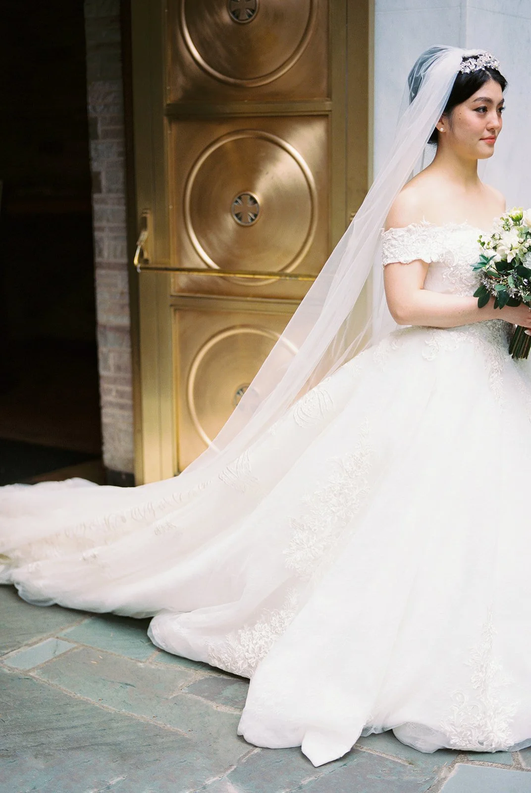 Full-length portrait of the bride in an off-shoulder lace ball gown with a long cathedral train and tiara, standing in profile before The Grotto's ornate gold doors