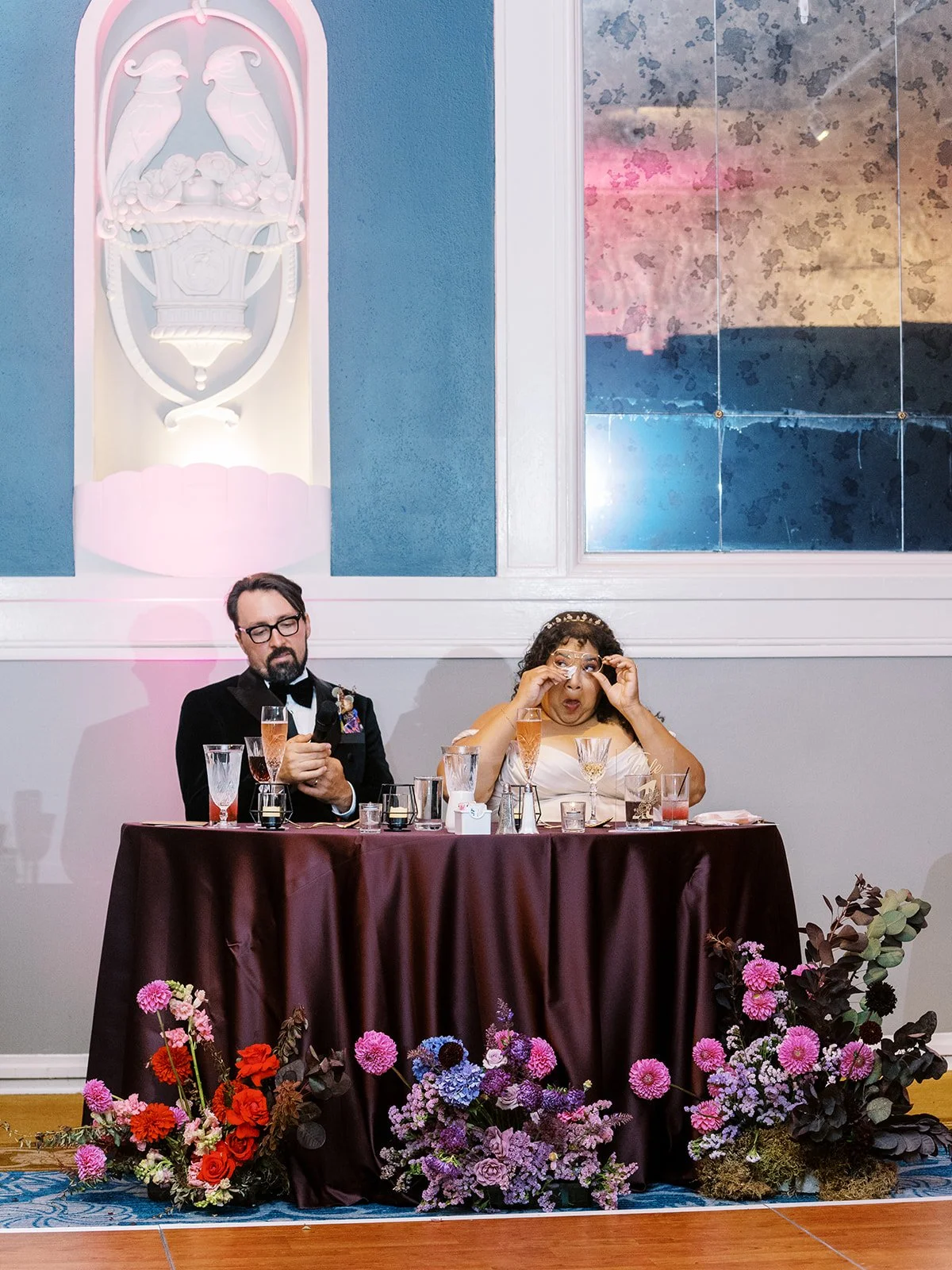 Couple seated at head table reacting during reception, surrounded by colorful floral arrangements and glassware.