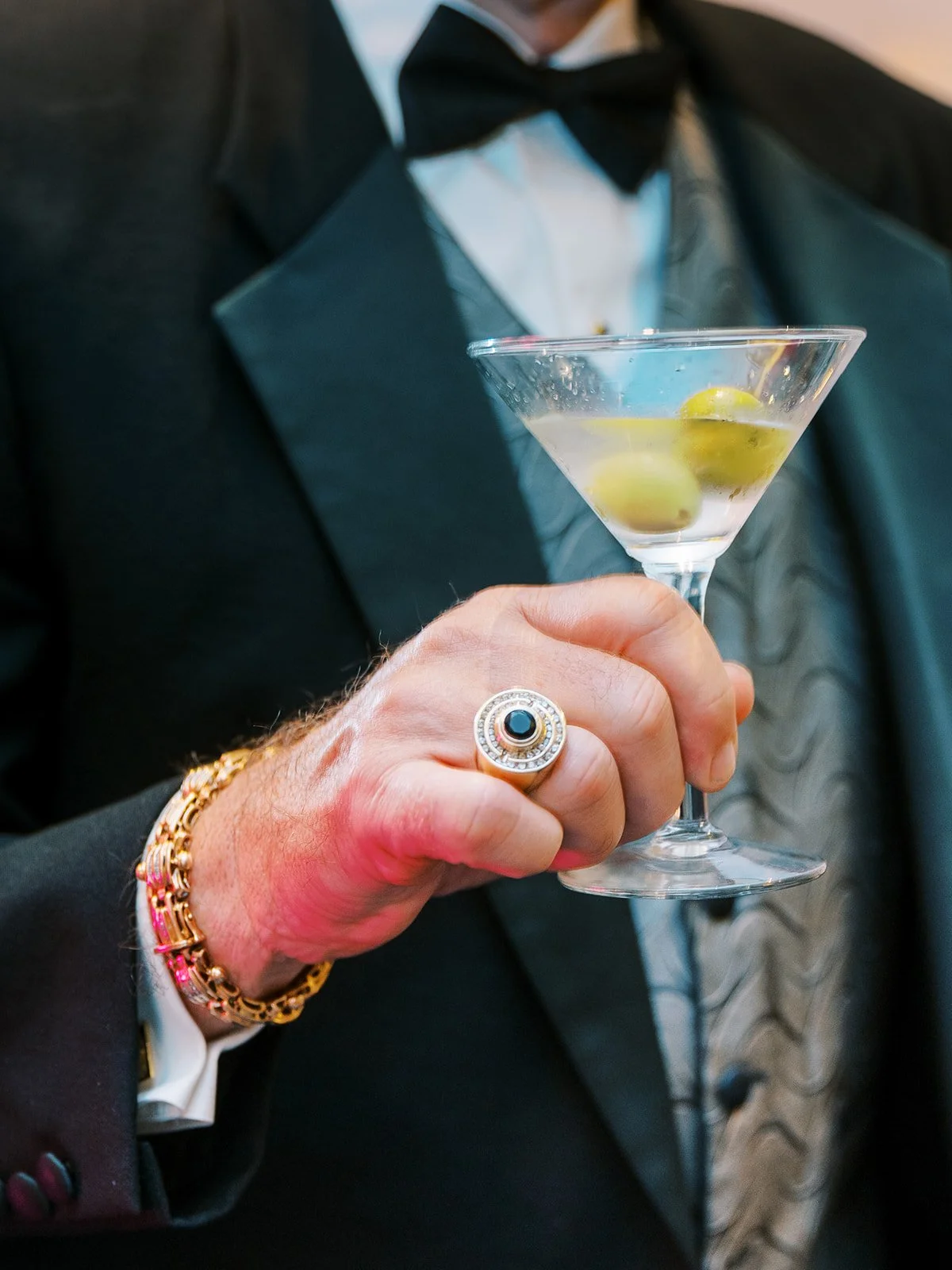 Hand in tuxedo holding a martini with olives, wearing a gold bracelet and ring, captured in soft event lighting.