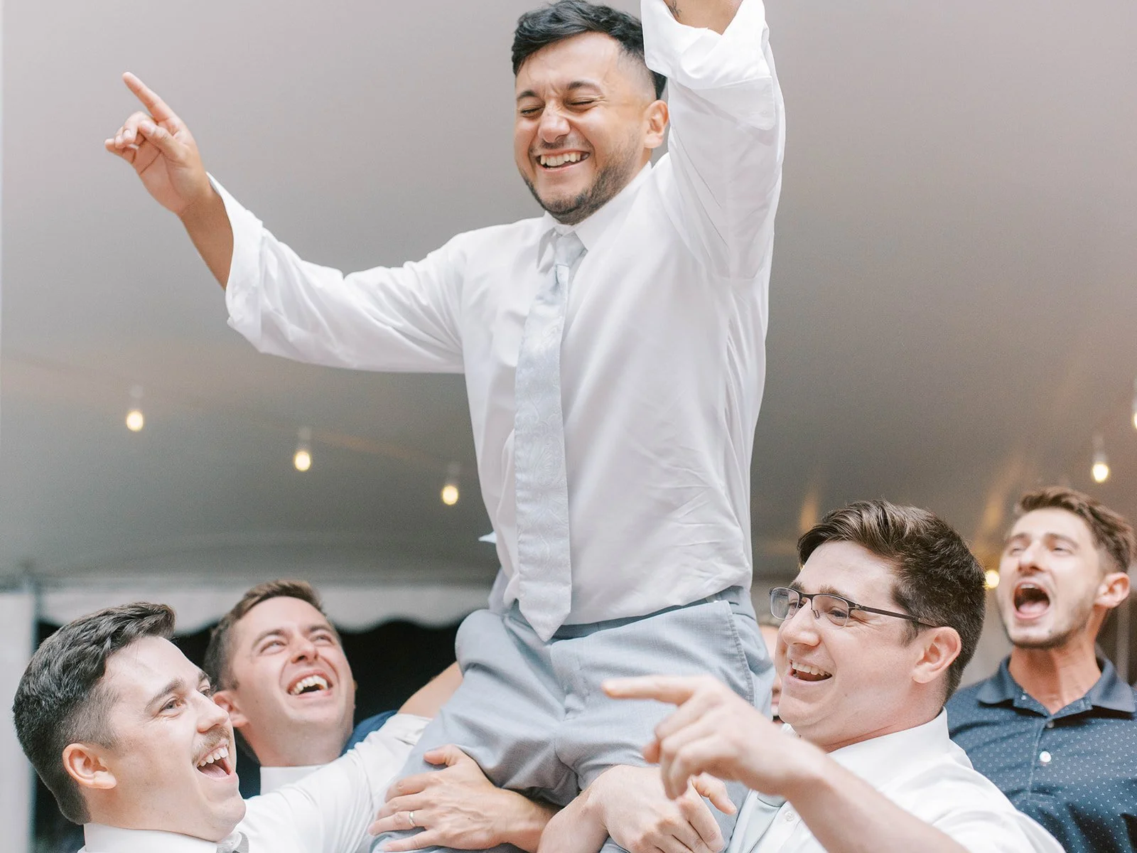 Groom smiles while lifted on guests’ shoulders during the wedding reception, surrounded by cheering friends.