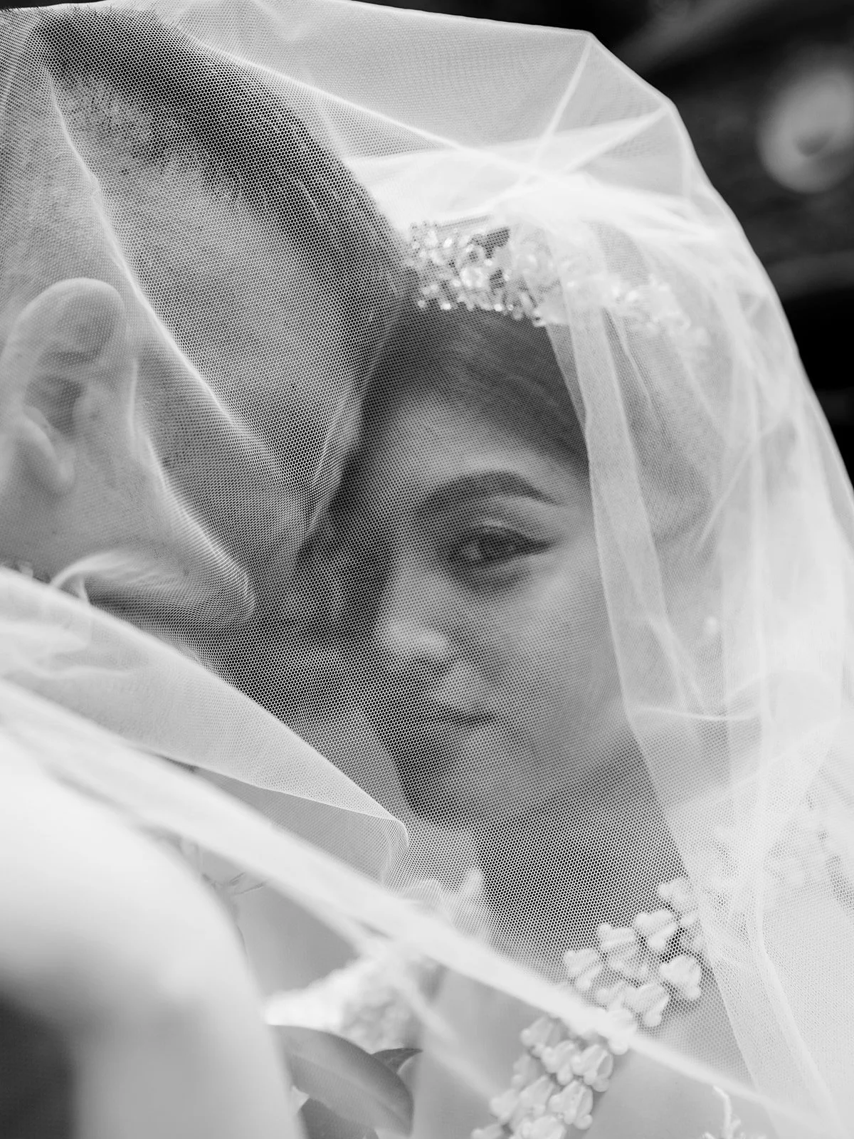 Black and white close-up of the bride's face partially obscured by her veil, her tiara and pearl earring visible through the tulle