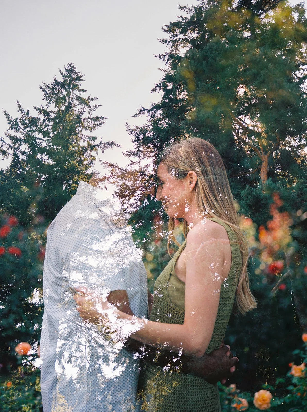 A dreamy double-exposure portrait of a couple facing each other, layered with evergreen trees and rose garden colors.