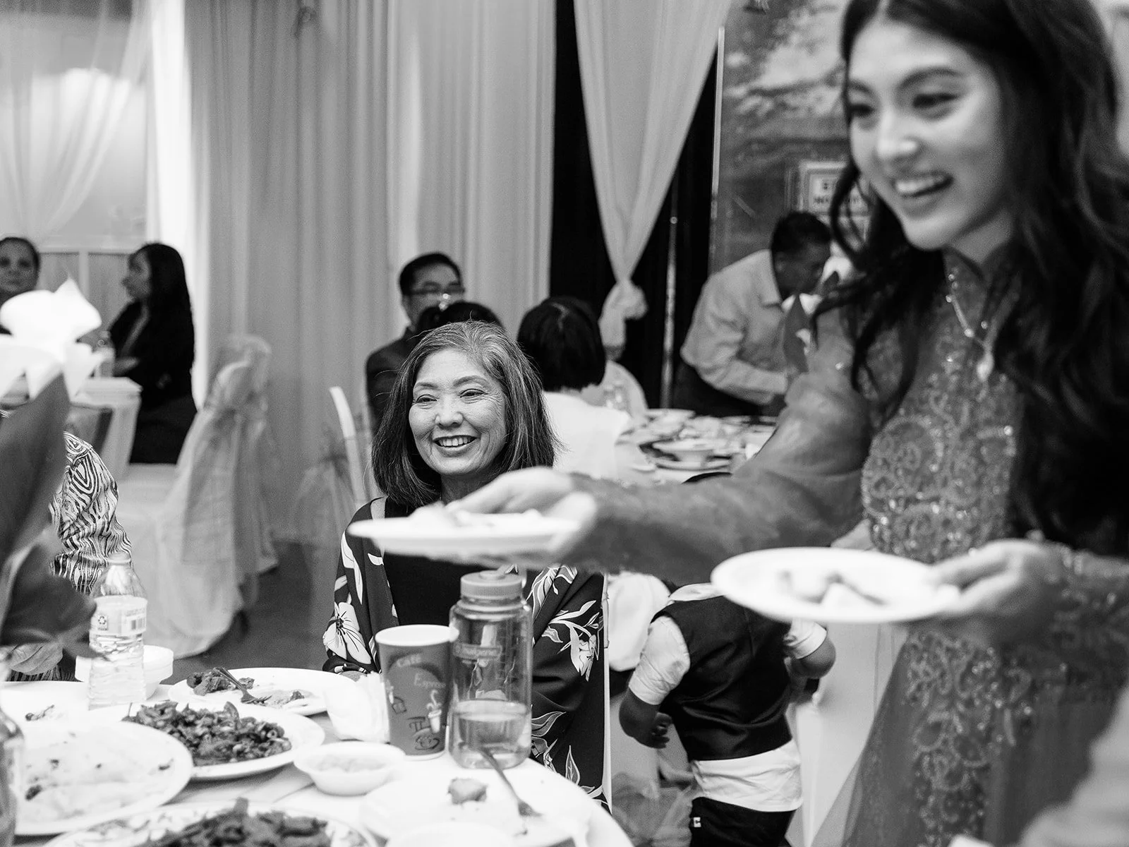 Black and white candid of a smiling older woman being handed a plate of cake at the Mekong Bistro reception, the bride visible laughing in the foreground