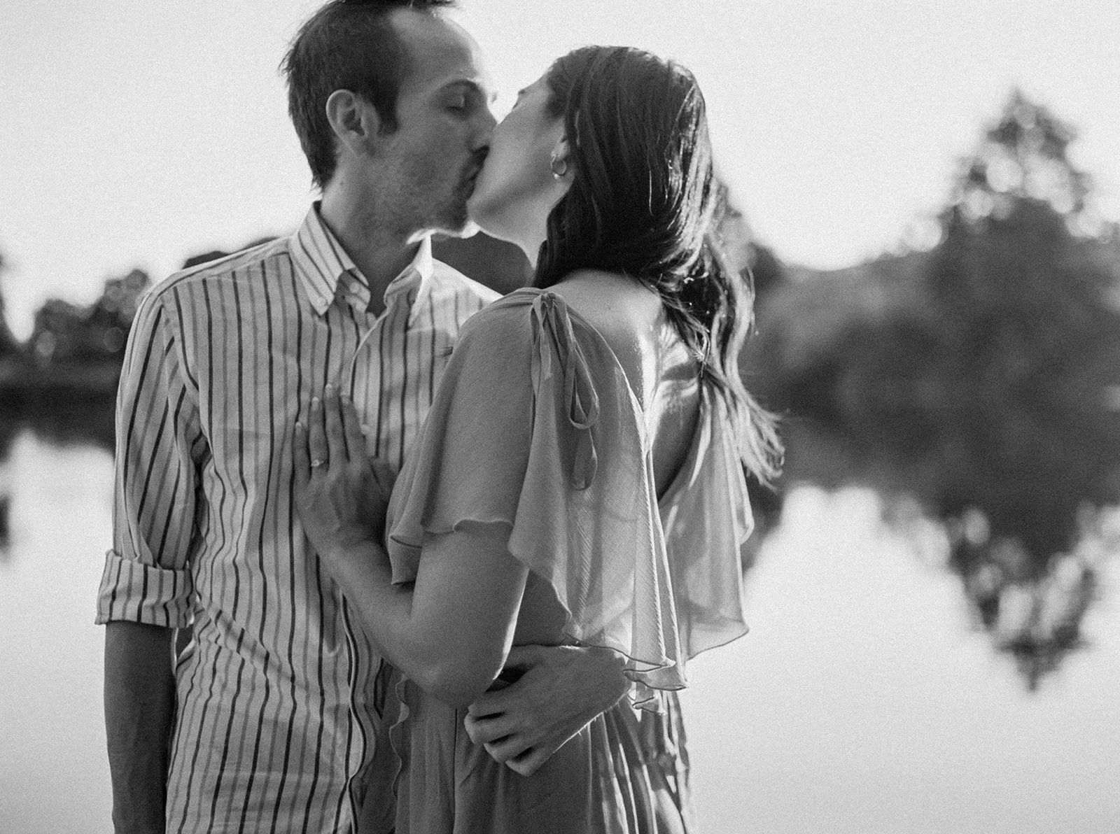 Black and white close-up of couple kissing by a lake, backlit by soft evening light during outdoor engagement session