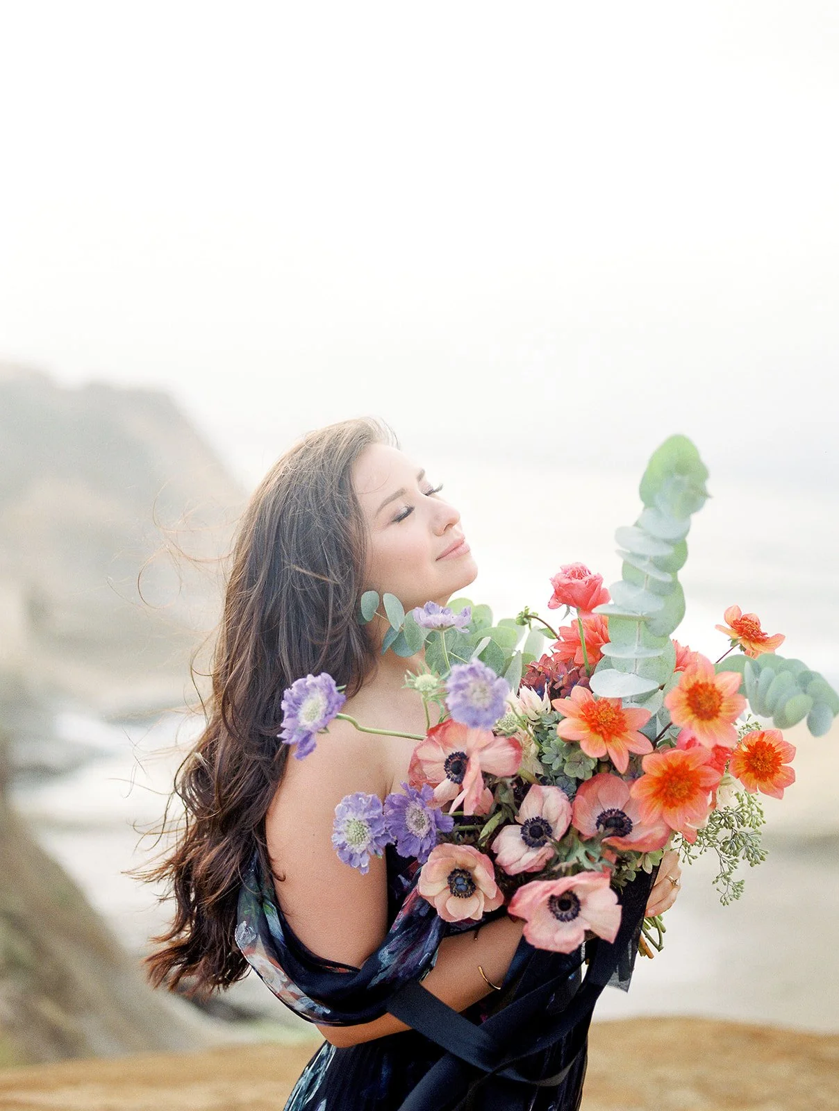 Woman holding colorful bouquet close, standing on coastal cliff with soft ocean background