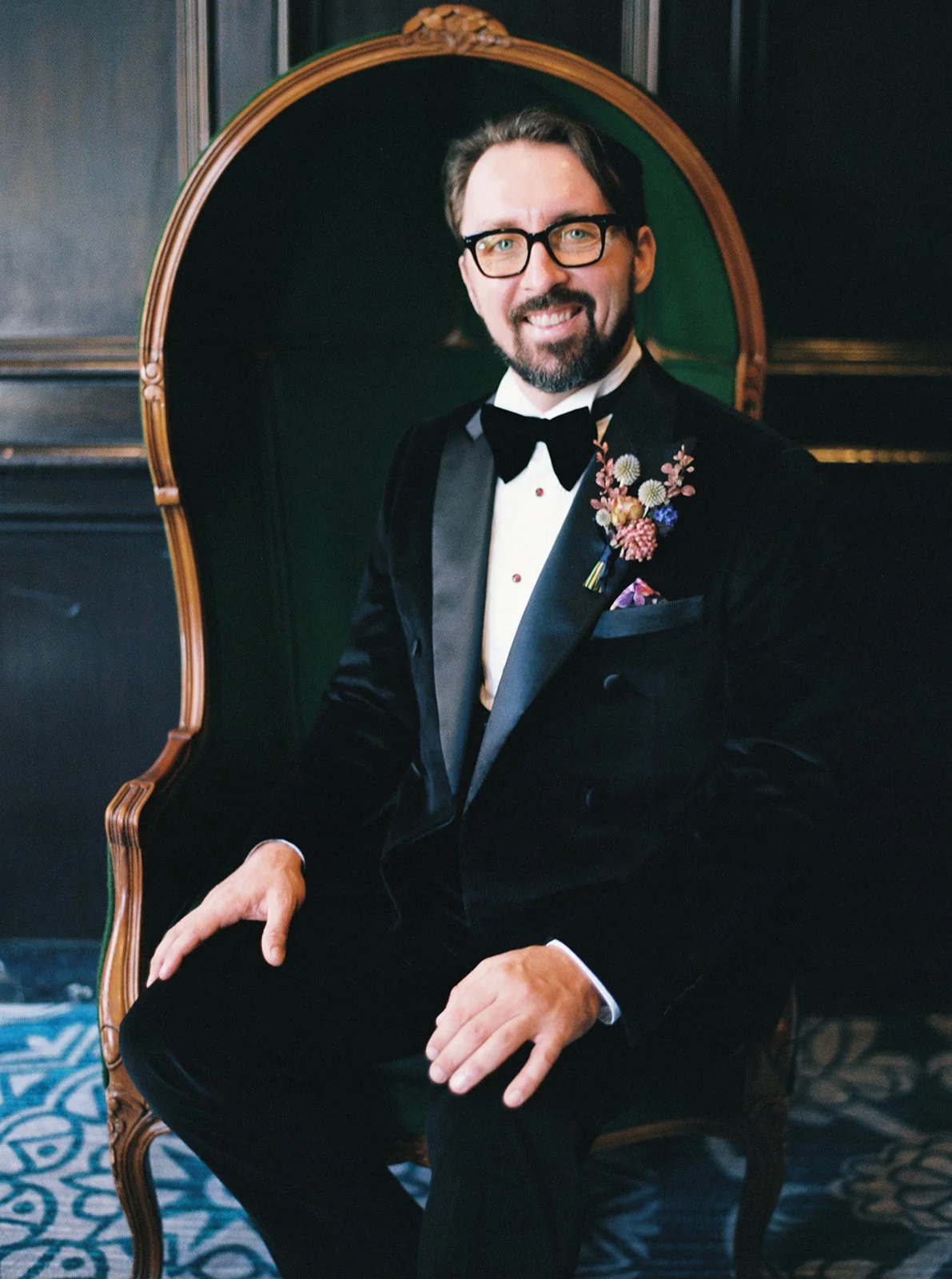 Groom in black tuxedo seated in a vintage chair, smiling, with boutonniere.