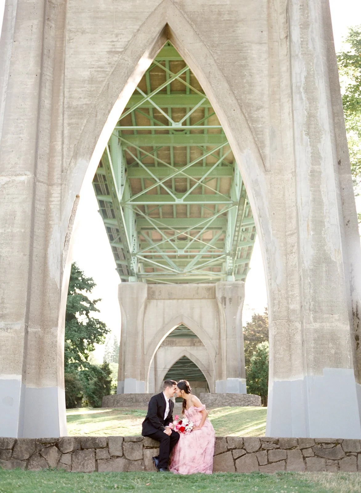 Couple sitting together beneath Cathedral Park bridge arches, framed by repeating columns.