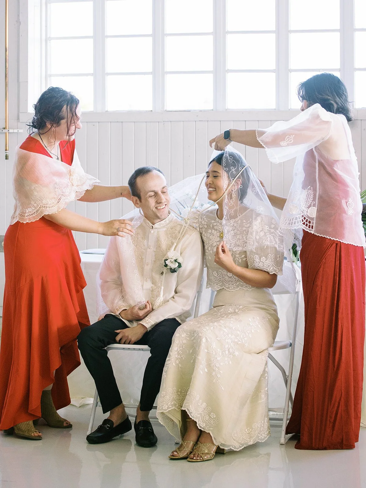 Two attendants in red Filipino attire placing the traditional wedding cord around the seated bride and groom during the ceremony
