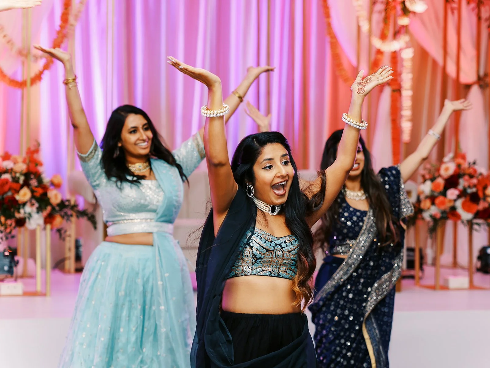 Bridesmaids in navy and pastel lehengas dancing in front of floral wedding backdrop.
