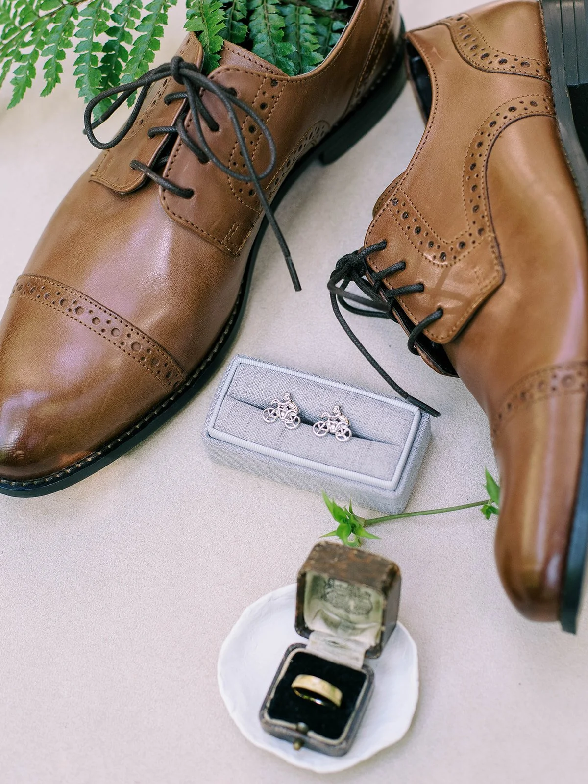 Brown leather dress shoes, cufflinks and a wedding band arranged with greenery.