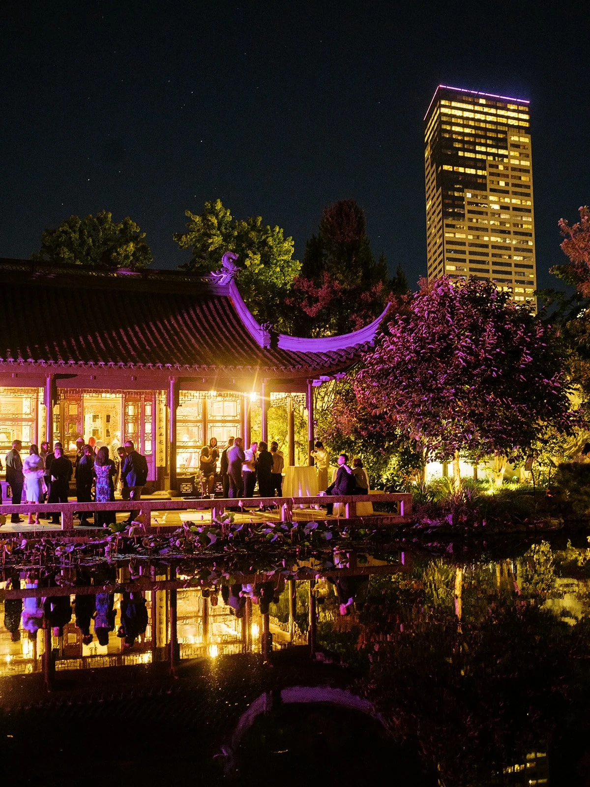 Wedding guests mingle on the pavilion terrace bathed in warm pink and gold uplighting at night, the glowing lattice screens and sweeping roofline reflected in the lake, with the Portland skyline rising behind.