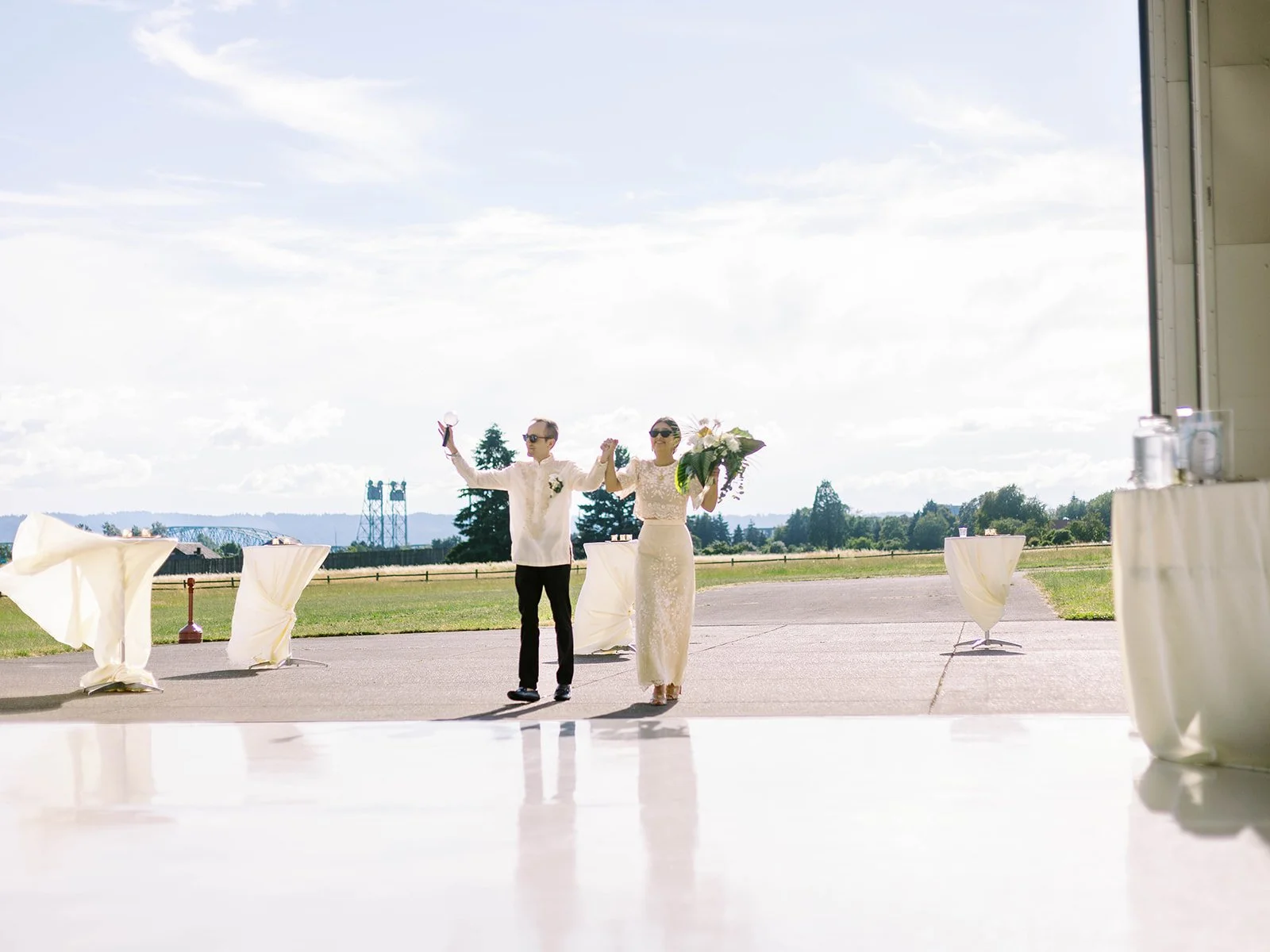 Wide shot of bride and groom in sunglasses raising their arms triumphantly as they make their reception entrance through the hangar doors
