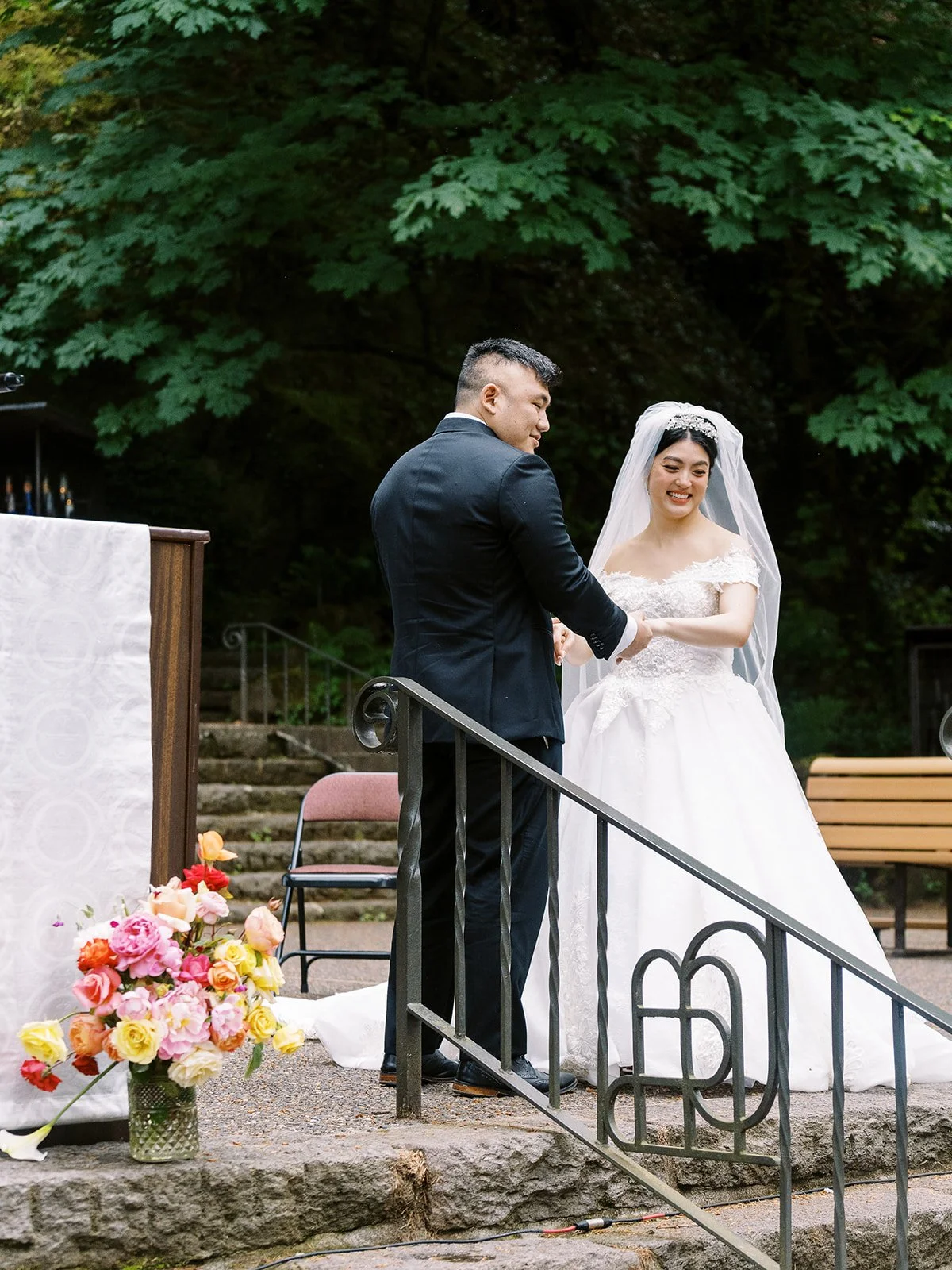 The groom takes the bride's hands and smiles at her at the altar, a colorful floral arrangement at the foot of the steps and forest greenery behind them at The Grotto