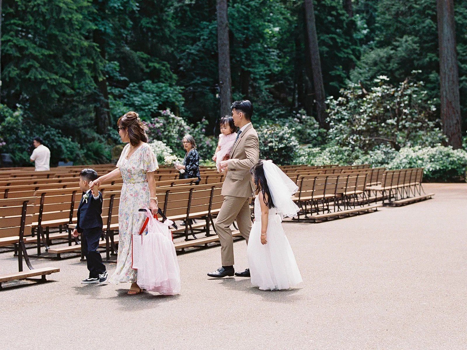 Wedding guests including a young ring bearer, flower girl in a white dress, and a man carrying a toddler walk through The Grotto's outdoor amphitheater seating before the ceremony