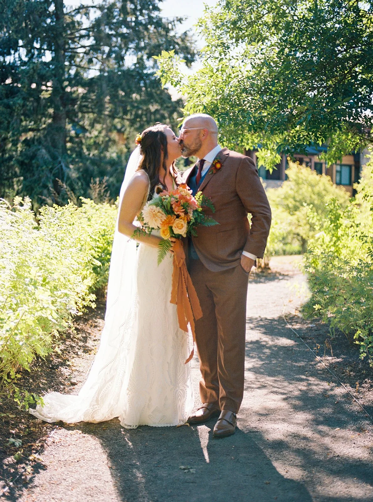 Bride and groom share a kiss on a sunlit garden path, holding a vibrant bouquet with warm-toned florals.