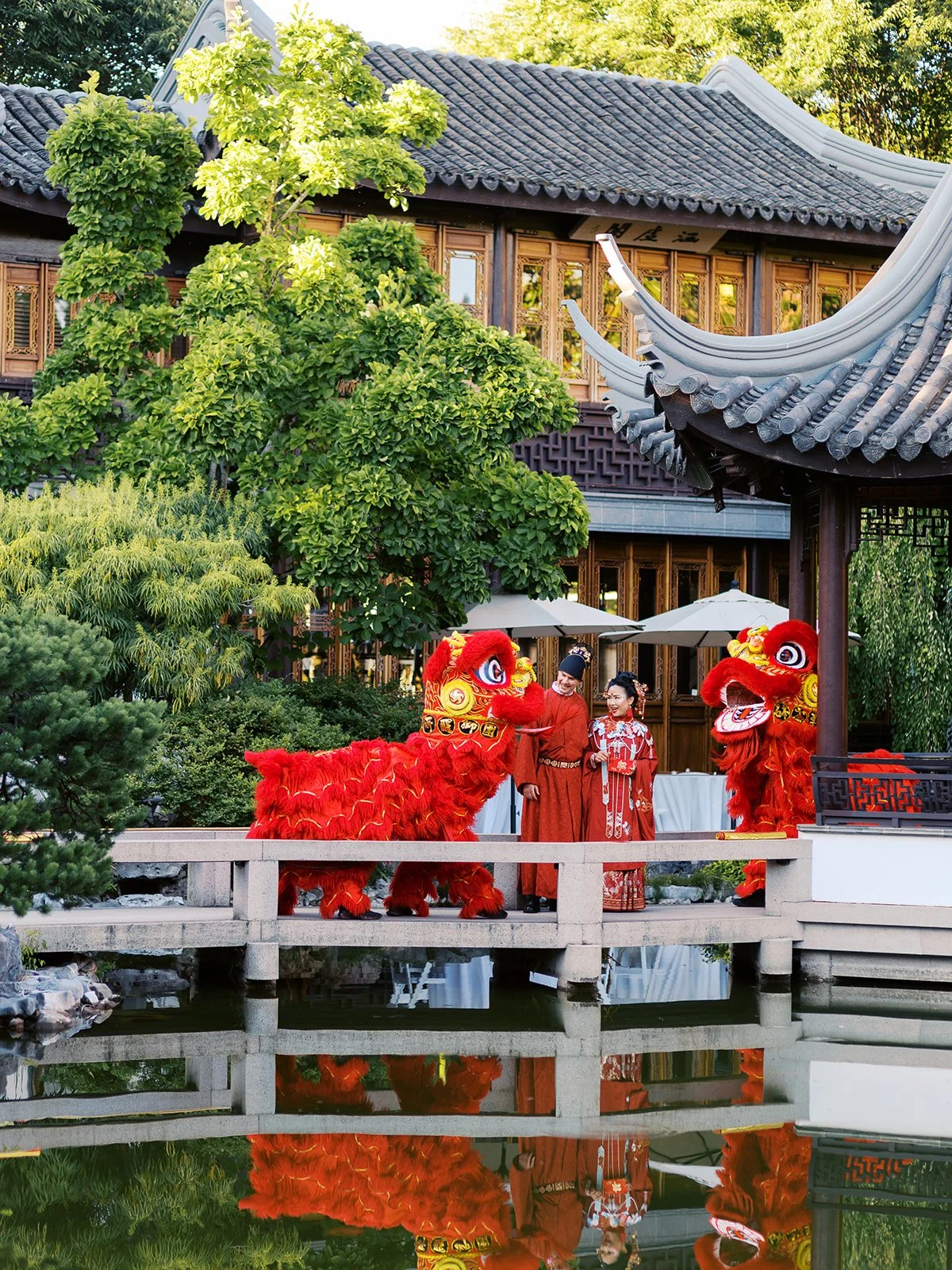 The couple in red traditional robes raise their hands in celebration on the stone bridge, flanked by attendants in hanfu and groomsmen in black suits, the garden reflected below.