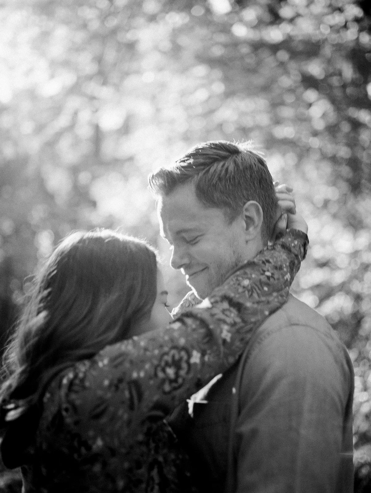 Close-up black and white portrait of couple embracing with soft light filtering through trees in forest